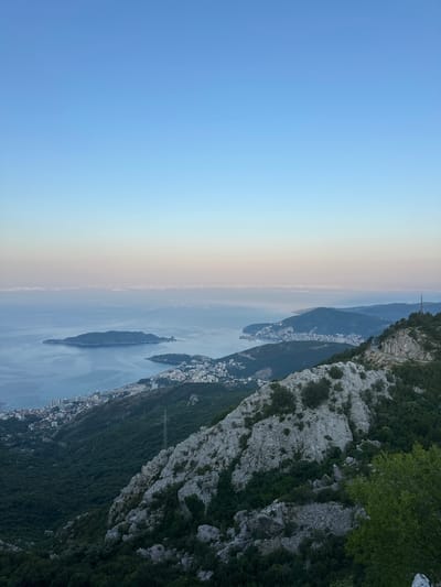 The image depicts a panoramic view from a mountainous vantage point overlooking the Adriatic Sea and the town of Brajici, Montenegro. The sky transitions from a clear blue at the top to a soft gradient of pink and light blue near the horizon, suggesting either sunrise or sunset. A thin band of clouds is visible along the horizon.
In the middle ground, the calm, blue sea stretches out, dotted with small islands and the coastline of Brajici. The town is characterized by white buildings nestled along the shore and climbing the hills. A prominent island, likely Sveti Nikola, is visible in the distance.
The foreground is dominated by a rugged, rocky mountainside covered in dense green vegetation. Large, light-colored rocks form part of the terrain, interspersed with trees and shrubs. A power line pylon stands on the slope, a stark industrial element amidst the natural landscape. The overall impression is one of a serene, natural environment meeting a coastal urban settlement, viewed from a significant elevation.