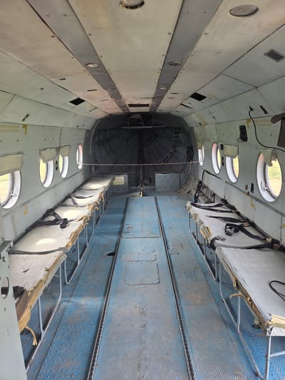The image shows the interior of a utilitarian aircraft, possibly a former medical evacuation aircraft, with rows of cots on either side of a central aisle. The cots are simple metal frames with worn, padded surfaces and black restraining straps. The floor is a durable blue metal with a patterned surface and a central channel. The walls are a light color, showing signs of age and some damage, with small circular windows spaced along the sides. The ceiling is also light-colored and features recessed lighting fixtures and ventilation grates. The rear of the cabin appears to be a large open cargo area with metal structures. Warning labels with Russian text are visible on the walls, including one that translates to "CUT OUT IN THIS AREA." The environment outside the windows appears to be a grassy, open field. The image does not contain any people or specific indicators of its exact location within Zhuchki, Russia.