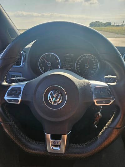 The image is a close-up, interior shot of a black leather steering wheel in a Volkswagen car. The steering wheel has silver accents and controls for the phone and audio system on the left side, and controls for the infotainment system and windshield wipers on the right. The Volkswagen logo is prominently displayed in the center of the steering wheel. The dashboard features a speedometer, tachometer, and a digital display showing the time, fuel consumption, and mileage. Outside the car, a rural landscape with windmills and trees is visible through the windshield under a bright, cloudy sky. The image appears to have been taken while the car was in motion. The location is identified as Meteren, Netherlands.