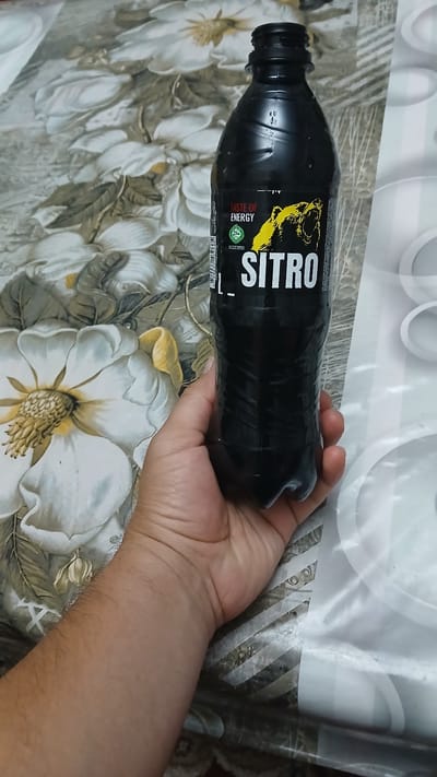 A hand holds a black plastic bottle of "SITRO" energy drink. The bottle has a yellow bear logo and the text "TASTE OF ENERGY" on its label. The background is a patterned tablecloth with floral motifs. The scene is indoors, on a table. There is no identifiable urban or geographical context visible to suggest a specific location in Kokand City, Uzbekistan.