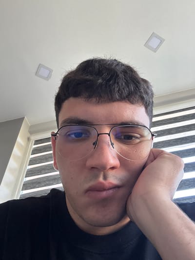 A close-up, low-angle, point-of-view selfie shows a man resting his head on his right hand. He is wearing black eyeglasses with thin metal frames and a black t-shirt. His dark hair is short and styled forward. The background features a white ceiling with two square light fixtures, and a window with horizontal blinds. The location is identified as Dibich, Bulgaria.