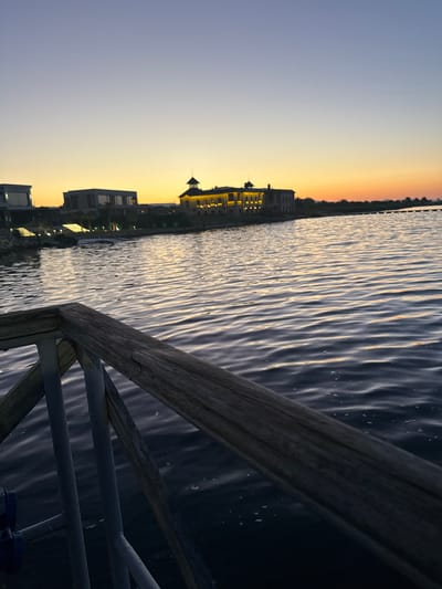 This is a photograph of a body of water at dusk, with buildings and a wooden railing in the foreground. The water is dark and appears rippled, reflecting the colors of the sunset. The sky transitions from a light blue at the top to a warm orange and yellow near the horizon. Several modern buildings are visible in the distance, some with illuminated windows. A prominent building with a tower and multiple windows is lit up, suggesting it is a significant structure. The wooden railing, made of weathered planks and metal supports, is in the lower left portion of the frame, angled diagonally. The scene appears to be a waterfront location. The prompt mentions Kazashko, Bulgaria, but there are no visual cues in the image to confirm this specific location.