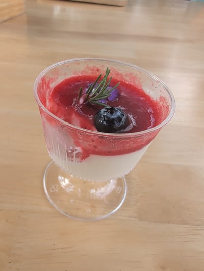 This media file is a top-down shot of a dessert served in a clear plastic cup. The dessert appears to be a layered confection, with a creamy white layer at the top and a vibrant red, possibly berry-based, layer below. A single dark blue blueberry sits on top of the red layer, and a sprig of rosemary with a small purple flower is artfully placed next to it. The cup is slightly tilted, and a clear plastic lid is resting beside it on a light-colored wooden surface. The wooden surface has visible grain and some small dark specks, suggesting it might be a table. The lighting appears bright, possibly indicating daytime, and there are no obvious weather cues. The setting seems to be an indoor or shaded outdoor area. No people are visible. The overall impression is one of a well-presented dessert. There is some faint embossed text on the plastic lid, possibly a logo or recycling symbol, but it is not clearly legible. The dessert is the central object of focus in this media file. The location context of Tambon Mae Raem, Thailand, does not provide any specific visual cues within the image itself.