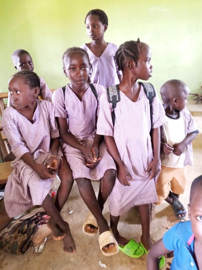 In Andaha, Nigeria, a group of young children, mostly girls wearing matching purple and white checkered school uniforms, are gathered. Some are seated on wooden benches or the floor, while others are standing. Several children are wearing backpacks. The environment appears to be a classroom or a similar educational setting, with a pale green wall in the background. The children's expressions vary, with some looking directly at the camera, others looking away, and some appearing thoughtful or curious. There are also a few stray items on the floor, such as sandals and a patterned bag.