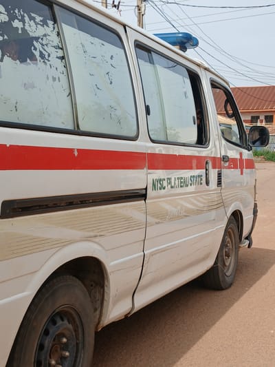 A white ambulance with a red stripe and "NYSC PLATEAU STATE" printed in green is parked on a reddish-brown road in Rayfield, Nigeria. A blue flashing light is visible on the roof of the vehicle. The ambulance's windows are dirty and partially obscured. In the background, a building with a brown roof and windows can be seen. Power lines are visible in the sky. The image is taken from a side-rear angle of the ambulance.