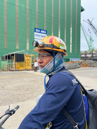 A man wearing a hard hat, safety glasses, and a blue long-sleeved shirt is shown in the foreground, riding a bicycle. The hard hat has a headlamp attached. The man has a blue and white patterned bandana covering the lower half of his face. He is wearing a backpack. In the background, a large green building with vertical ribbed siding is visible, along with a sign that reads "2C4 AREA". There are also cranes and stacks of metal scaffolding. The scene appears to be an industrial area, possibly a shipyard, in Geoje-si, South Korea. A yellow container with a radiation symbol is also visible.