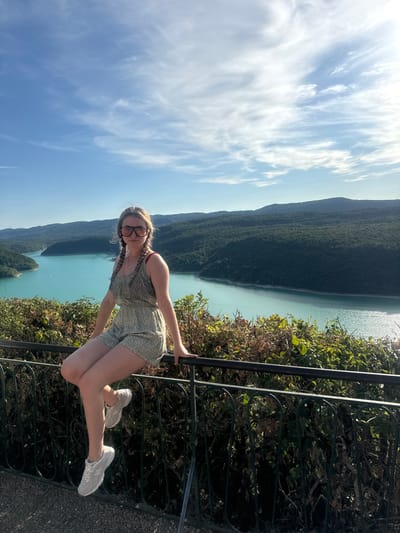 A young Caucasian woman with blonde hair in braids and sunglasses is seated on a dark metal railing. She is wearing a floral print romper and white sneakers. She is looking at the camera. Behind her, a body of turquoise water, likely a lake or river, is surrounded by lush green, tree-covered hills. The sky is blue with scattered white clouds. The scene appears to be an overlook, possibly in Moirans-en-Montagne, France, known for its scenic beauty and natural landscapes.
