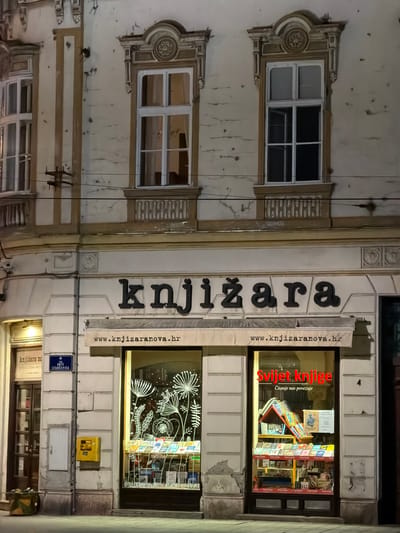Here's a description of the image:
The image depicts the facade of a building at night. The building houses a bookstore (knjižara in Croatian). The bookstore is called "Knjižara Nova." Two large windows display books and decorative floral graphics. "Svijet Knjige" is visible in one window, with the tagline "Čitanje nas povezuje". A sign below the main bookstore sign includes the bookstore's website: www.knjizaranova.hr. A blue street sign indicates the location as "Ante Starčevića", a street name in Osijek, Croatia. A small yellow post box is mounted near the street sign. Above the bookstore front are windows with decorative frames and signs of aging/disrepair on the building's facade. Electrical wires are visible across the building's front. A smaller sign reads "knjižara no SVEUČILIŠNA KNJIZ - STUDELJISKI CD - DVD AUDIO."