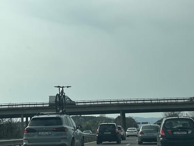 The image shows a traffic jam on a highway near Lesichovo, Bulgaria. Several cars are visible, including a gray BMW SUV with a bicycle mounted on its roof, a black Toyota RAV4, a gray BMW sedan, and a black SUV. License plates are visible on some of the cars. An overpass spans the highway. The sky is overcast. The environment includes trees, hills, and a highway with multiple lanes.