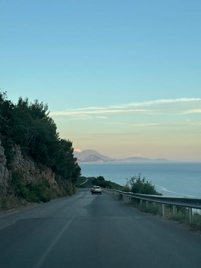 The image shows a winding road along a coastline in Višnjevo, Montenegro, with a car driving ahead. The road curves to the right, bordered by a steep, rocky embankment covered in dense green foliage on the left. A metal guardrail runs along the right side of the road, separating it from a vast expanse of blue sea. In the distance, rolling hills and mountains are visible, with a hazy, pastel-colored sky above, suggesting either sunrise or sunset. The sea appears calm, with a few wispy clouds in the sky. The car in front is white and appears to be a hatchback or SUV. The overall scene conveys a sense of travel and scenic beauty, characteristic of coastal roads.