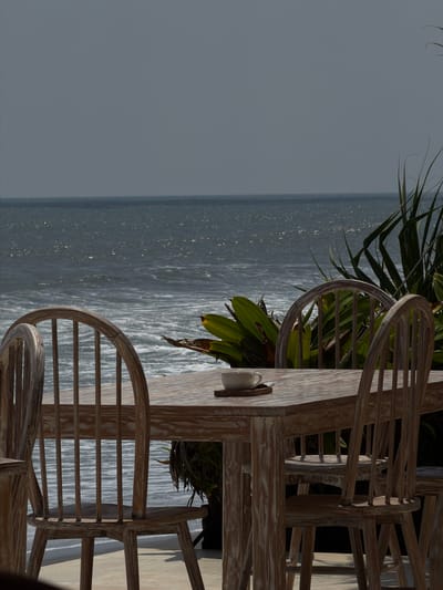 An outdoor seating area in Belalang, Indonesia, features a weathered, light-colored wooden rectangular table with four matching spindle-back chairs, two prominently facing the ocean. A white ceramic cup rests on a dark brown coaster on the table surface. Beyond the seating, a dark blue-gray ocean with white-capped waves occupies the mid-ground, extending to a hazy gray horizon beneath an overcast sky. Lush, spiky green tropical foliage partially frames the ocean view from the right and center-right. The ground beneath the table and chairs is a light-colored, flat surface. No individuals are present in the scene.
