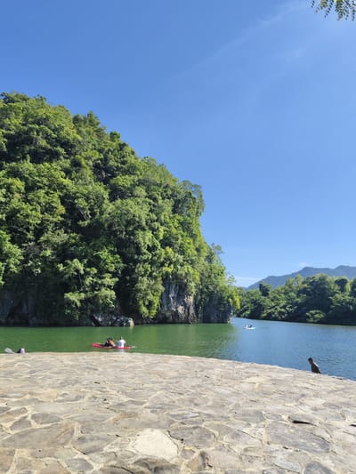 The image captures a serene natural landscape on a bright, clear day, likely in Lamseunia, Indonesia. The foreground is dominated by a textured stone pathway, laid with irregular, flat stones, suggesting a man-made structure bordering a body of water.

To the left, a lush, green, tree-covered cliff face rises dramatically from the water's edge. The water itself is a vibrant emerald green, reflecting the surrounding foliage and the clear blue sky. In the middle distance, two red kayaks are visible on the water. One kayak has two individuals, one appearing to be paddling, and the other seated. The second kayak is closer to the shore and has one person visible. Further out on the water, a third, smaller light-blue boat with at least one person is also present.

The background transitions to rolling hills covered in dense green vegetation, creating a layered effect of natural beauty. The sky is an uninterrupted, deep blue, indicative of fair weather. On the stone pathway in the foreground, a single dark-colored figure, possibly a person, is perched on the edge, looking out towards the water. Another figure is partially visible on the far left of the stone path, near the water. The overall impression is one of a peaceful recreational area where visitors can enjoy the natural environment.