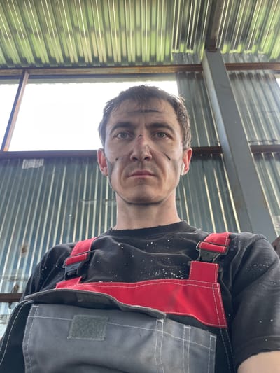 A low-angle, waist-up selfie shows a man with dirt smudges on his face and chest. He is wearing a black t-shirt with white speckles and red and gray overalls. The man has short, brown hair and is looking directly at the camera with a neutral expression. He has a white earbud in his right ear. Behind him, a corrugated metal wall and ceiling are visible, with a large window on the left side of the frame. The overall setting appears to be an industrial or workshop environment. The lighting suggests it is daytime, possibly outdoors or in a well-lit indoor space. The image was taken in Sokury, Russia.