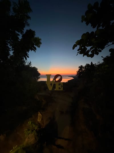 The image depicts a sunset scene in Karimunjawa, Indonesia, featuring large yellow letters spelling out "LOVE" in the foreground, positioned on a rocky path. The letters are illuminated, with the "O" appearing red. Behind the letters, a body of water stretches to the horizon, where the sky transitions from a deep blue at the top to vibrant orange and pink hues near the water's surface, indicative of a sunset. Silhouettes of trees frame the scene on both the left and right sides, with some leaves visible in the upper right portion. In the distance, the faint outlines of islands or landmasses can be seen. The overall lighting suggests it is either dusk or dawn.