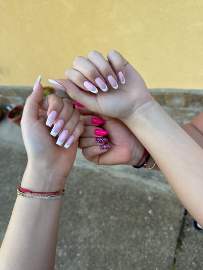 The image shows two pairs of arms and hands. One pair has long, squared-off nails with a classic French manicure. The other pair has shorter, pointed nails, some painted a bright pink and others with a black and pink leopard print design. Both wrists wear bracelets. The background is blurry but appears to be an outdoor setting, with what looks like a yellow wall and a concrete surface, possibly a patio or sidewalk. The location is Lehchevo, Bulgaria.