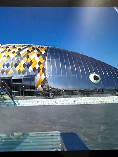 This media file features a close-up, vertically oriented shot of a building designed to resemble a fish. The building's head, which faces upwards in the frame, is a metallic blue with horizontal lines suggesting scales. A large, round eye with a black pupil and a light green iris is prominent on the head. Below the head, the building's body is covered in overlapping, scale-like tiles in a pattern of yellow, gold, white, and dark blue.
The building appears to have windows and access points, with visible metal walkways and railings attached to its side. To the right of the fish-shaped building, there is a textured, grey surface, possibly another structure or a wall, with a blue band running across it. Further to the right, a corrugated metal structure is visible.
The background is a bright, clear blue sky, suggesting a daytime scene with fair weather. No people are clearly discernible in the image. The overall impression is that of an unusual architectural structure, likely an art installation or a uniquely designed building in an outdoor setting. The location context provided is Lopatino, Russia. There is no visible text in the image.