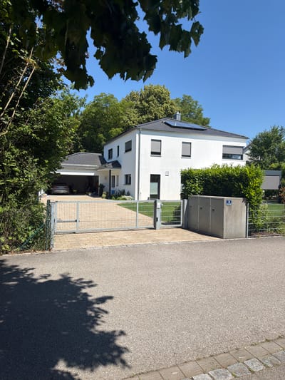 The image shows a modern, two-story white house with a dark gray tiled roof and solar panels. The house has several windows with closed gray roller shutters. A double garage is attached to the left side of the house, and a car is parked inside. The property is enclosed by a light gray metal fence with a gate. In front of the gate, a paved driveway leads to the garage. Adjacent to the fence on the right side of the property are three gray concrete utility boxes, one of which has a blue number "3" sign. A dense green hedge runs along the front of the utility boxes and continues around the property. Lush green trees surround the house, and the sky is clear blue. The scene appears to be a residential street in Ingolstadt, Germany, judging by the architectural style and the presence of elements common in German residential areas. The foreground shows the asphalt road with shadows cast by trees.