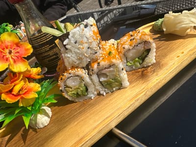 The image shows a plate of sushi on a wooden platter. The sushi consists of several rolls with fillings, including what appears to be shrimp and avocado. Some of the sushi is coated in sesame seeds, and others in orange roe. Accompanying the sushi are garnishes of wasabi and pickled ginger. A decorative plastic flower arrangement sits to the left of the sushi rolls, along with a small bottle of soy sauce. The platter rests on a black table, with glimpses of outdoor seating and a bicycle in the background.
Given the German text on the soy sauce bottle, it is highly plausible that the picture was taken in Germany. However, there is no clear evidence to place the picture specifically in Rotenburg an der Fulda, Germany.