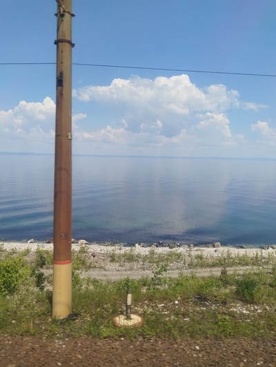 This image depicts a serene natural landscape adjacent to the city of Solzan, Russia. The scene is dominated by a vast expanse of calm, blue water, likely Lake Baikal, reflecting the clear blue sky dotted with fluffy white clouds. Along the shoreline, a pebbly beach transitions into a grassy embankment dotted with small green bushes. In the foreground, a tall, weathered utility pole stands prominently, its surface showing signs of age and use, with a visible number "30" towards its base. This pole is likely part of the infrastructure supporting the railway line, as indicated by the tracks and ballast visible at the very bottom of the frame. The overall impression is one of peaceful natural beauty, with human-made elements integrated into the environment. There are no people or animals visible in the frame.