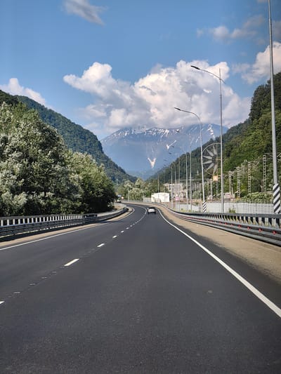 The image depicts a highway winding through a mountainous landscape, likely in or near Chvizhepse, Russia, under a bright blue sky with scattered white clouds. On the left, a dense green forest with a large tree covered in white blossoms lines the side of the road. The highway has multiple lanes, indicated by white lines, and is bordered by a metal guardrail with reflective elements. A single white car is visible in the distance on the highway. On the right side of the road, there are streetlights, utility poles, and what appears to be railway infrastructure. In the background, a majestic mountain range with snow-capped peaks dominates the horizon, contrasting with the lush green slopes of the surrounding mountains.