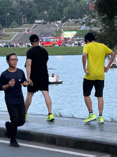 In Xuan Huong Lake, Da Lat, Vietnam, three individuals are visible. In the foreground, a person wearing glasses and a dark long-sleeved shirt and black pants is jogging. To their right, a person in a black t-shirt and black shorts stands facing away from the camera. Further to the right, a person in a bright yellow t-shirt and black shorts is also facing away. In the background, a red and orange bus is parked near a grassy slope with stairs, where many people are gathered. The lake is calm with a small white boat in the middle. Trees and distant buildings are visible on the far shore.