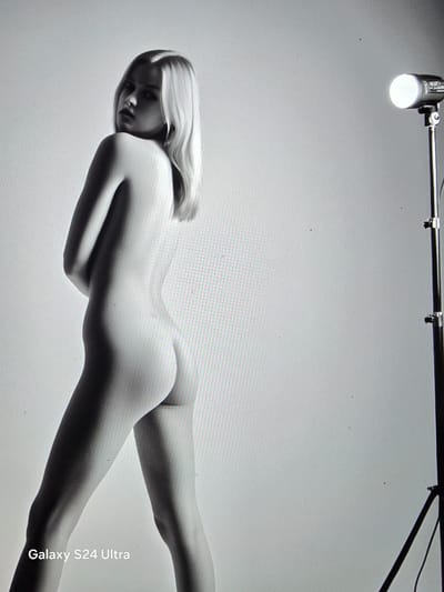 A nude young woman with blonde hair is posing with her back to the camera, looking over her shoulder. She is standing in a studio with a white backdrop and a lighting rig to her right. The image is a black and white photograph. The location is unknown.