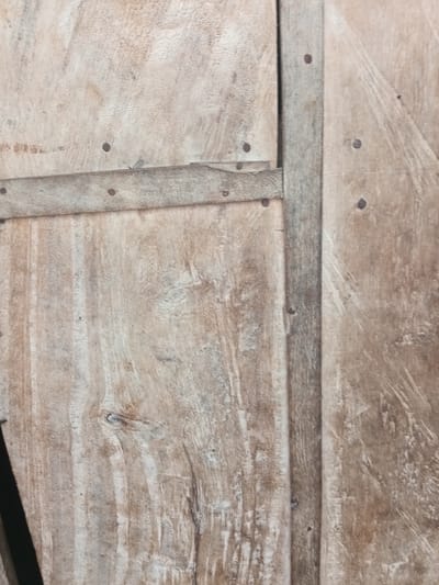 The image shows a close-up view of a weathered wooden surface, likely the side of a crate or a rustic door. The wood appears to be composed of several planks joined together. There are visible nails holding the planks and reinforcing strips in place. The texture of the wood is rough and aged, with prominent grain patterns and some discoloration, suggesting exposure to the elements. The overall impression is one of age and durability. The image does not contain any visual information that would indicate its location in Zinna, Nigeria, or any other specific city. There are no people, actions, or interactions depicted.