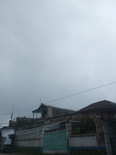 The image depicts a static scene within the city of Eheliyagoda, Sri Lanka, under an overcast sky. In the foreground, a low, light-colored concrete wall with a blue base stripe and weathered texture runs horizontally. A green, vertical-bar metal gate is centrally located within this wall. Behind the wall, to the left, a multi-story structure is visible, featuring a lower section partially covered by a white tarp, possibly indicative of a temporary shelter or stall. Above this, the structure appears to be concrete, with exposed rebar and an unfinished or dilapidated upper level characterized by a skeletal pitched roof frame and visible interior elements. A flagpole, bearing a red and white horizontal striped flag, extends upwards from this building's left side. To the right of the central wall, a building with a traditional reddish-brown tiled roof is partially visible, obscured by dense green foliage. Overhead, multiple black utility lines traverse the frame horizontally. The ground level in the immediate foreground is unpaved, appearing as dirt or rough terrain with minimal vegetation. No individuals or active movements are observed.