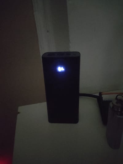 A black power bank is positioned on a light-colored surface, possibly a table or shelf. A blue digital display on the power bank shows "64". To the right of the power bank, a dark cable extends from it and connects to a black charging adapter, which is partially visible. Next to the adapter, a cylindrical metallic object with a cap is standing upright, appearing to be a water bottle or flask. The background consists of a wall with a textured surface. The lighting is dim, suggesting an indoor environment in low light conditions. There is no information about the location in the city of Kipchak, Tajikistan.