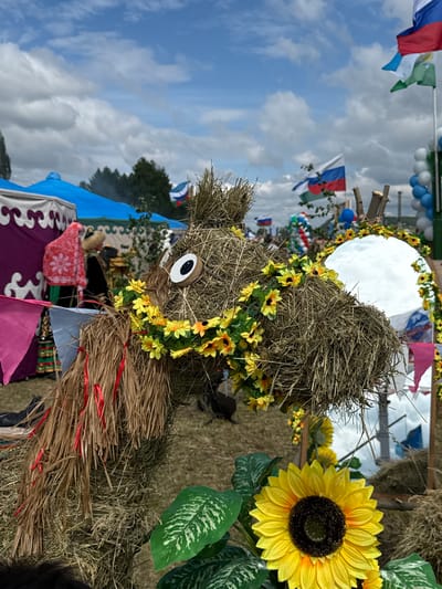 This is a medium-shot, eye-level, outdoor photograph taken on a cloudy day. The foreground is dominated by a straw sculpture of an animal, possibly a camel or horse, adorned with a garland of yellow artificial sunflowers and red ribbons. The sculpture has a large, cartoonish eye made of wood and black felt. To the right of the sculpture is a round mirror reflecting the sky and surrounding scene, framed by straw. In front of the mirror, a large, artificial sunflower is placed.

In the middle ground, there are several tents with colorful fabrics, including a purple and white patterned yurt-style tent and a blue tent. People are visible in traditional clothing, one wearing a red headscarf and a decorated outfit, and another in darker clothing with a fur-trimmed hat. Russian flags are visible on flagpoles in the background. Balloons in white, blue, and green are also present, suggesting a festive occasion. The ground is covered in dry grass and straw.

The background shows more tents, trees, and the sky with scattered clouds. The overall impression is of an outdoor festival or fair held in a rural or semi-rural setting, potentially in or near the village of Старое Субханкулово, Russia, given the Russian flags and cultural elements.