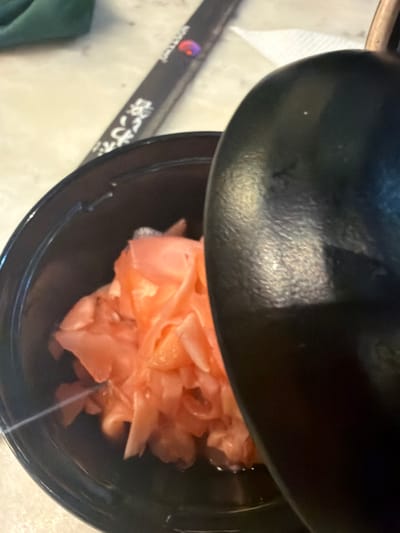 A portion of pickled ginger, its pale pink color prominent, sits in a black bowl, partially obscured by a black, round lid. The ginger appears to be thinly sliced and piled loosely. Beside the bowl, a pair of chopsticks with Japanese writing rests on a light surface. The scene is shot from a top-down perspective, giving focus to the ginger and surrounding tableware.