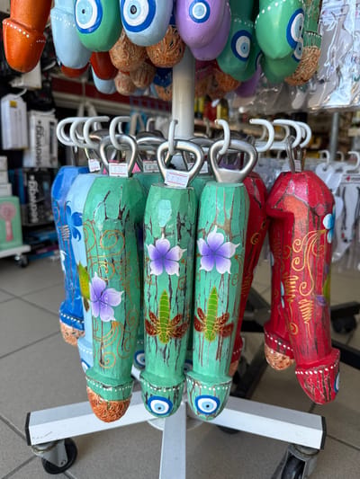 In Agía Marína, Greece, a close-up shot captures a display of colorful, phallic-shaped trinkets hanging from a white metal rack. The trinkets are made of wood and feature hand-painted designs. Several are green, adorned with floral motifs and golden swirls, while others are red and blue, some decorated with "evil eye" symbols. The trinkets have metal loops at the top for hanging. In the background, shelves filled with various souvenirs and merchandise are visible, suggesting a retail environment, likely a souvenir shop. The overall scene is bright and captures a detailed view of these distinctive decorative items.