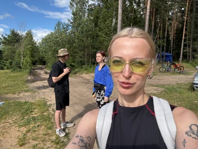 This is a selfie taken by a young woman with blonde hair and yellow sunglasses. She is wearing a black tank top with white and red accents. She has tattoos on her left arm.

In the background, three other people are visible. To her left, a person in a blue jacket is seen from behind. To her right, a man wearing a black t-shirt and black shorts is standing. Further in the background, another person, possibly male, is wearing a camouflage hat and a black shirt.

The scene appears to be outdoors, likely in a park or a natural area, with trees and a dirt path. There is also a bicycle visible in the middle left of the image. The sky is blue with scattered clouds, suggesting it's daytime with pleasant weather.

The orientation of the photo is unusual, rotated 90 degrees clockwise. The subjects are looking at the camera, and the overall mood seems casual and relaxed, perhaps a group outing or a day spent outdoors. There is no visible text in the image.

The location context is Desnogorsk, Russia.