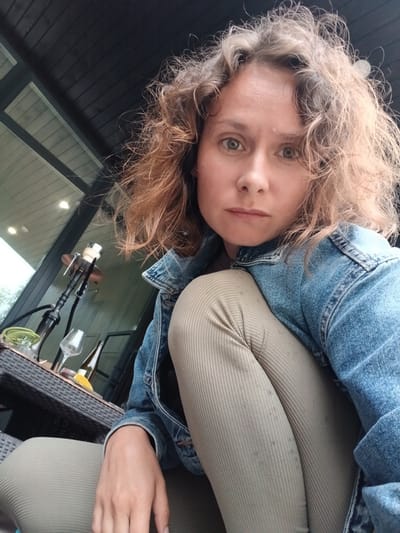 A young woman with curly, light brown hair is captured in a selfie, taken outdoors in what appears to be a backyard or patio setting. She is wearing a denim jacket over a beige ribbed top and pants. Her head is tilted, and she is looking directly at the camera with a relaxed expression.

In the background, a dark, slatted wooden structure, possibly part of a house or fence, is visible. To her right, on a dark wicker table, are several objects: a hookah, a wine glass, and a bottle, suggesting a leisurely activity. The scene is lit by natural light, indicating it is daytime, with the sun casting gentle highlights on her hair and skin. The overall mood is casual and relaxed. The location is Verkhneye Dubrovo, Russia.
