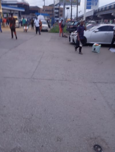 The image depicts a street scene in Port Harcourt, Nigeria, likely in the evening or overcast conditions given the lighting. Several individuals are visible on a paved road. In the foreground, a person in a striped shirt and dark pants walks towards the right, holding a phone to their ear. To their left, a light blue bag with an orange handle sits on the ground. Further in the background, a person in a patterned outfit walks from left to right, followed by another person in a white shirt and dark pants. A white car is parked on the right side of the road. In the mid-ground, to the left, a person in a green shirt stands near a vehicle, and a cart with a rider is visible. The background shows buildings and overpasses, typical of an urban environment. The overall scene suggests a bustling public space with pedestrian and vehicular traffic.