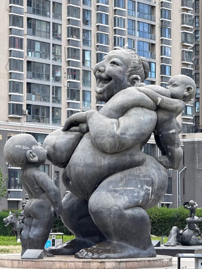 The image depicts a bronze sculpture of a woman carrying three children on her back and shoulders. The sculpture is located in Changping District, China. The background features a tall building with multiple windows. In the vicinity of the sculpture, there is a patch of green grass, a bush, and other bronze sculptures, including one of an animal. The sky is overcast, casting soft lighting on the scene. The sculptures appear to be in an outdoor urban setting.
