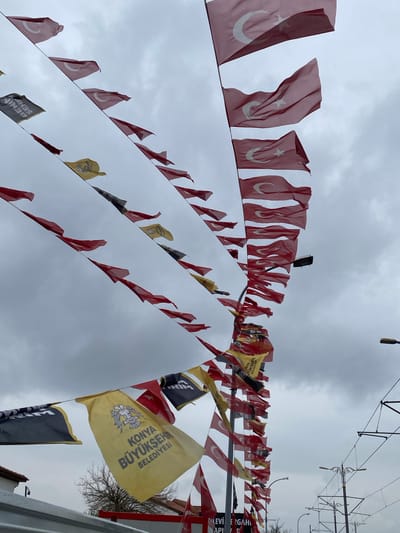 The image shows a low-angle view of multiple rows of flags strung across an overcast sky. The flags are of two main types: Turkish flags, red with a white crescent and star, and yellow and black flags. The yellow flags are printed with the words "KONYA BÜYÜKŞEHİR BELEDİYESİ" and feature an emblem. Some black flags are also visible. Power lines and associated hardware can be seen in the upper right. The scene is likely located in Karatay, Turkey, suggested by the text on the yellow flags, which refers to the Konya Metropolitan Municipality.