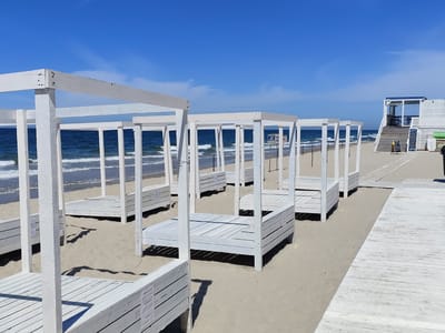 The image shows a sunny day at the beach in Yantarny, Russia. The beach is lined with white wooden structures, likely sun loungers or cabanas, arranged in rows on the sand. The sea is visible in the background with gentle waves. There are wooden boardwalks on the sand, and a white building with blue accents and stairs leading up to it is visible on the right side of the image. No people are present in the scene. The sky is a clear blue with a few wispy clouds.