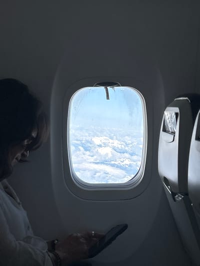 A woman is seated by an airplane window, looking out at a view of clouds and blue sky. Her head and upper body are visible in the lower portion of the frame. She holds a dark-colored object in her right hand, raised near the window. She is wearing bracelets and a ring. The interior of the airplane is visible, including the window frame, reading light, and overhead console. The location is not determinable from the image.