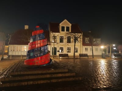 The image captures a night scene in Hans Jensens Stræde, Odense, Denmark. A prominent conical spiral sculpture, approximately 3-4 meters tall, stands centrally on a three-tiered base. The sculpture is constructed from alternating red and dark blue rectangular ceramic tiles arranged in a helical pattern. The ground, paved with cobblestones, is wet and highly reflective, suggesting recent precipitation, with streetlights casting bright glares. In the background, a row of traditional two-story buildings with pitched red tile roofs is visible. The central building is light-colored with dark window frames and a gabled dormer. Bare deciduous trees are sparsely distributed in front of the buildings, and at least four dark-colored park benches are present. Multiple streetlights illuminate the scene, and a silver public waste receptacle is visible on the right side of the frame. No individuals are present, and no discernable actions are taking place.