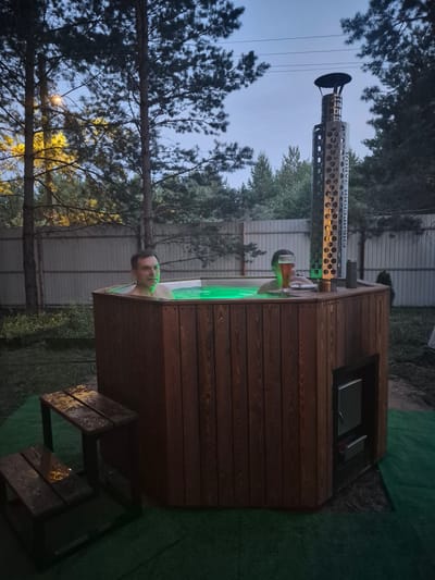 In Staraya Binaradka, Russia, at dusk, two men are relaxing in a wooden hot tub illuminated by green LED lights. The hot tub is situated outdoors in a grassy area, surrounded by trees and a fence. A tall metal chimney with hexagonal cutouts stands beside the hot tub, and a glass of beer rests on the edge of the tub. A small wooden stool is placed next to the hot tub, with water droplets visible on its surface. The sky is a dusky blue, with the silhouette of trees against it.