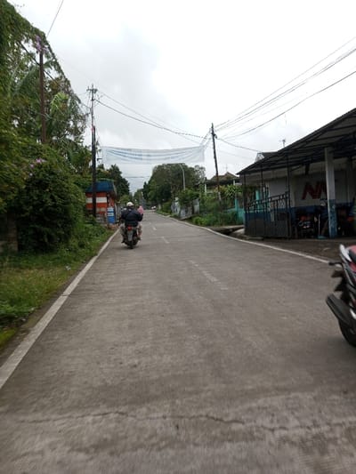 The scene depicts a paved road in Melakudikadu, India, under an overcast sky. The concrete road surface features visible cracks and a white line marking its left edge. A dark-colored motorcycle, operated by an individual wearing a dark jacket and light head covering, travels away from the viewer in the left half of the roadway. A pink item is visible behind the rider. On the left side of the road, dense green foliage, including vines, covers a utility pole and partially conceals a small structure with red and white vertical stripes. A white banner is suspended horizontally across the road above this section. On the right, a commercial building with a corrugated metal roof protrudes towards the road. Its facade displays a blue section with an 'MM' logo, and several figures are discernible near it. The front wheel and engine area of a red and black motorcycle are partially visible in the immediate foreground on the right. The road extends into the background, flanked by utility poles with multiple overhead wires, additional vegetation, and various structures. A second white banner is visible spanning the road further in the distance.