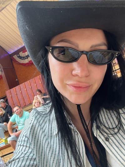 A close-up, selfie-style shot shows a woman wearing a black cowboy hat and black cat-eye sunglasses. Her dark hair is visible around her face. She is wearing a striped button-up shirt. In the background, out of focus, are several other people sitting on wooden benches. The environment appears to be an indoor venue with wooden paneling and a large American flag decoration. The lighting suggests it is daytime. The scene is likely a social gathering or event.