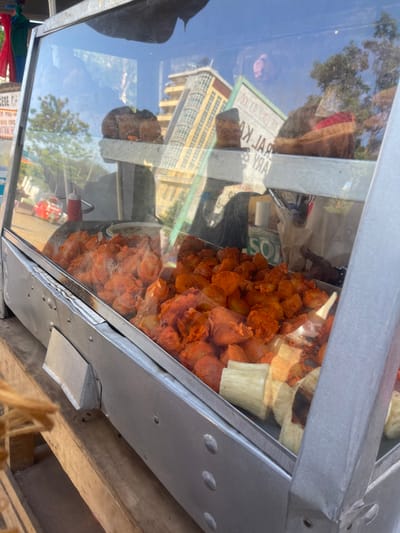 The image, rotated 90 degrees clockwise, shows a close-up of a glass and metal food display case, likely part of a street vendor's cart. Inside, a generous portion of reddish-orange, irregularly shaped fried food is visible, possibly savory snacks or fried dough. Next to these, a few lighter-colored, rectangular food items, resembling tamales or wrapped pastries, are stacked.
The glass surface of the display case strongly reflects the outdoor surroundings. A bright blue sky with green tree foliage is clearly seen. Also reflected is a multi-story building with yellow walls and a pattern of arched windows. Partially visible and inverted text on a reflected sign can be deciphered as "KAL KAA" followed by "J R Y" or "ARY". A small red object, possibly a fire extinguisher, is also reflected near the bottom.
The bright, natural lighting indicates it is daytime. The scene suggests a commercial food service setting, likely outdoors in an urban or semi-urban area, consistent with a street food vendor. Although the context mentions a "Hospital ward, Kenya," the visual evidence points to an external environment, perhaps a street or market near a hospital, rather than an indoor ward. No people are directly visible, but the setup implies an active vending operation.