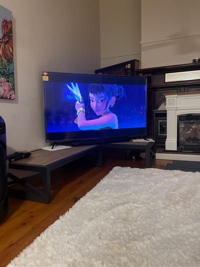 The image depicts an indoor living room scene. A large flat-screen television displays an animated character with dark hair styled in buns, holding a glowing blue weapon. The television is positioned on a dark wooden coffee table with a metal frame. To the left of the television, a portion of a painting with a floral motif is visible on the wall. In the foreground, a plush white rug covers a section of the wooden floor. To the right of the television, a fireplace with a white mantelpiece and a dark insert is present, flanked by a built-in cabinet. The overall ambiance suggests a comfortable, modern living space. The image does not contain information to suggest it is located in Bendigo, Australia.