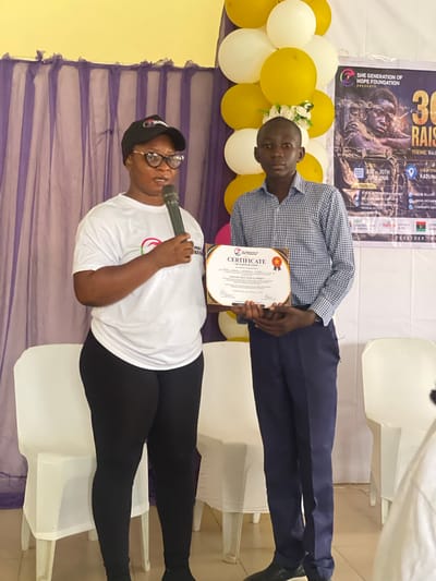 Two people are posing for a photo in front of a backdrop. The person on the left is a woman wearing glasses, a black baseball cap, a white t-shirt, and black leggings. She is holding a microphone and smiling. The person on the right is a man wearing a checkered blue and white shirt and dark blue pants. He is holding a certificate. In the background, there is a banner with text that includes "SHE GENERATION OF HOPE FOUNDATION PRESENTS 30 RAIS" and "THEME: Rec", "DATE: 1ST-30TH JUNE 2026", and "LOCATION: KADUNA". There are also balloons in yellow and white. The scene appears to be an indoor event. The overall impression is one of celebration or recognition, possibly an award ceremony or a community event held in Kaduna, Nigeria.