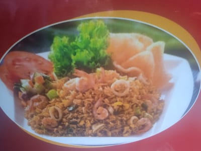 The image is a close-up of a plate of fried rice. The fried rice is a light brown color and appears to contain small pieces of shrimp and squid rings. It is garnished with lettuce, sliced tomatoes, and a few crackers. The plate is white and rests on a red and yellow background. There is no information about the location in the city of Peurupok, Indonesia.