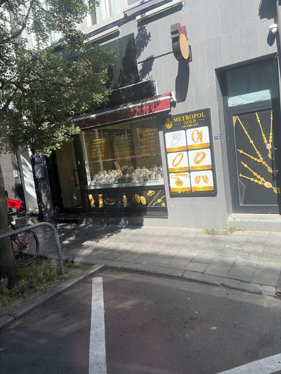 This is a medium-shot, high-angle photograph of a jewelry store called "Metropol Gold" located in Saint-Josse-ten-Noode, Belgium. The store is situated on a street with a sidewalk paved with square tiles and an asphalt road marked with a white line. The building facade is gray and the store has a large glass display window where jewelry is showcased. Above the window, there's a decorative golden chain motif. To the right of the entrance, there's a sign with the store's name and various jewelry items like rings, bracelets, and necklaces. The sign also indicates the gold purity: "14K-18K-22K" and displays Turkish words for different jewelry types: "Alyans" (ring), "Bilezik" (bracelet), "Pırlanta" (diamond), and "Kolye" (necklace). The number "72" is visible on a blue sign near the store.
In front of the store, a tree with dense green foliage casts shadows on the sidewalk. A bicycle is parked to the right of the image, near a metal railing. The scene appears to be set during the daytime, with bright sunlight casting distinct shadows, suggesting a clear and sunny weather. There are no people visible in the image, but the presence of a bicycle and the store's display indicate it is likely an active commercial area. The overall impression is that of a typical street view of a jewelry shop in an urban setting.
