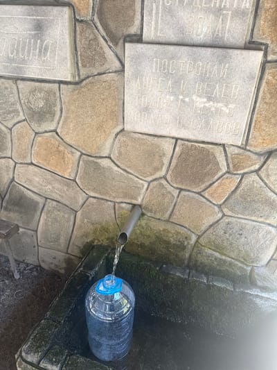 The image depicts a spring in the city of Ravnogor, Bulgaria. A stone wall, constructed with irregularly shaped stones of varying colors, forms the backdrop. A stone plaque is embedded in the wall, bearing engraved text in what appears to be Cyrillic script. Below the plaque, a metal pipe protrudes from the wall, from which clear water is flowing into a large, transparent plastic bottle with a blue cap. The bottle is positioned in a stone basin, part of the spring's structure, which appears wet and mossy. To the left of the spring, a wooden bench is partially visible. The overall scene suggests a functional water source, likely a public fountain or natural spring, maintained with a rustic aesthetic.