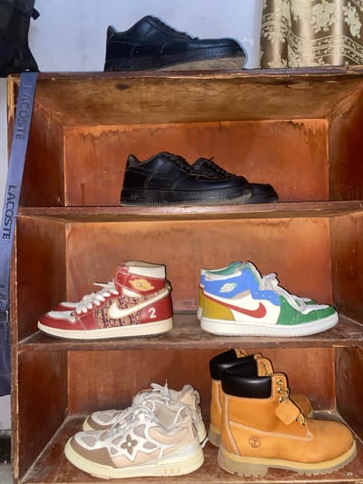 The image captures a collection of six pairs of sneakers and boots meticulously stored within a rustic, multi-compartment wooden shelf unit. No individuals are present in the scene.
From left to right and top to bottom:
The top left shows a partial view of a black Nike Air Force 1 shoe hanging from the shelf edge. Below it, a pair of black Nike Air Force 1 sneakers rests within a compartment.
The top middle compartment features a single, colorful high-top sneaker, resembling a customized Nike Air Jordan 1. It displays a prominent red Nike swoosh against panels of green, blue, yellow, and white.
To the top right, a pair of classic tan Timberland boots with black padded collars and the distinctive Timberland tree logo is visible.
The bottom middle compartment holds a single red and beige high-top sneaker. This shoe exhibits a patterned design, with visible "Dior" branding and the number "2" on its side, suggesting a designer collaboration or inspired piece.
In the bottom right, a pair of beige and white Louis Vuitton Trainer sneakers is displayed, featuring their characteristic multi-paneled design and quatrefoil motifs.
A dark blue strap, clearly labeled with the text "LACOSTE" in white, spans horizontally across the bottom of the image, beneath the shoe rack. A patterned curtain or fabric is partially visible in the top left background. The wooden shelf unit appears aged, with a visible grain and some signs of wear, indicating a functional storage setup in an indoor setting. The scene reflects organized storage of various branded footwear. The location context is Jama A, Nigeria.